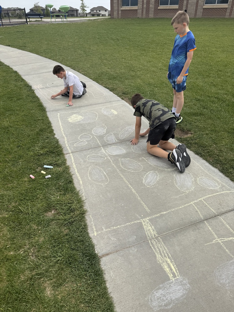 Three young boys are working with sidewalk chalk on a concrete path next to a grassy field. Two boys are kneeling, drawing large circles and rectangular borders with yellow and white chalk, while a third boy in a blue athletic shirt stands by watching. Several pieces of colorful chalk lie in the grass nearby.