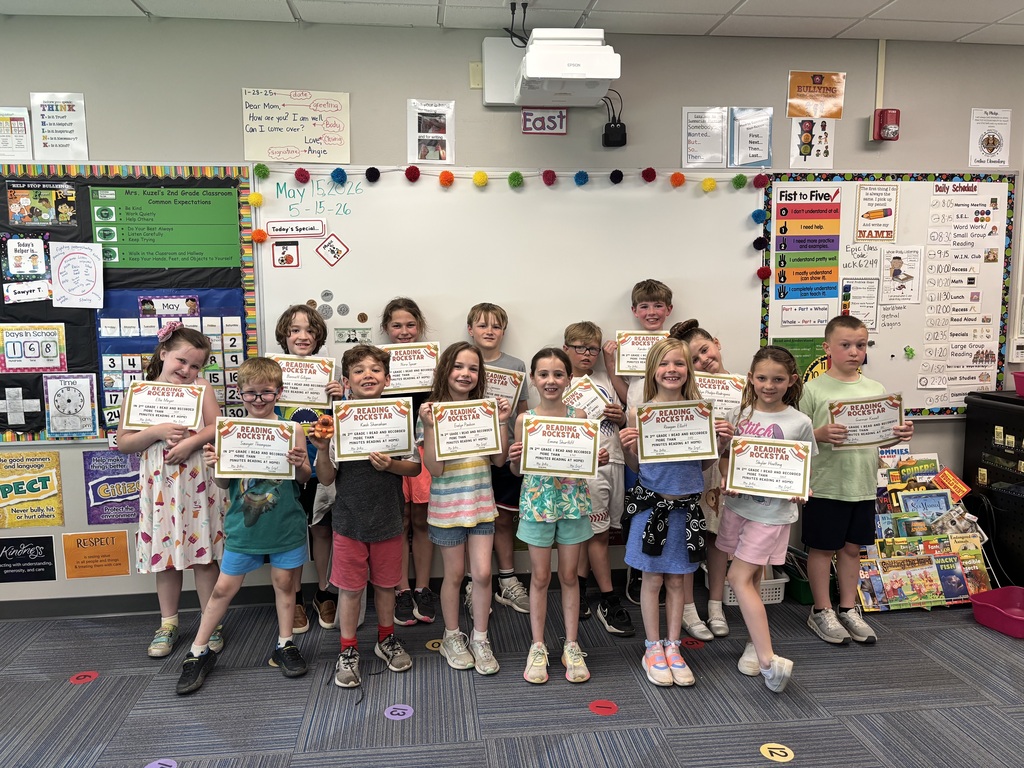 A classroom group photo of thirteen second-grade students standing in front of a whiteboard, proudly holding "Reading Rockstar" certificates. The room is decorated with educational posters, a daily schedule, and a "Fist to Five" chart. The date "May 15, 2026" is written on the whiteboard above the children.