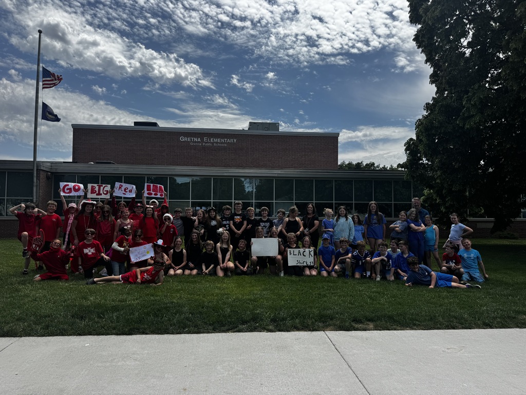 A large group of elementary students wearing red, black, and blue clothing pose together for a group photo on a grassy lawn in front of Gretna Elementary School under a cloudy sky.