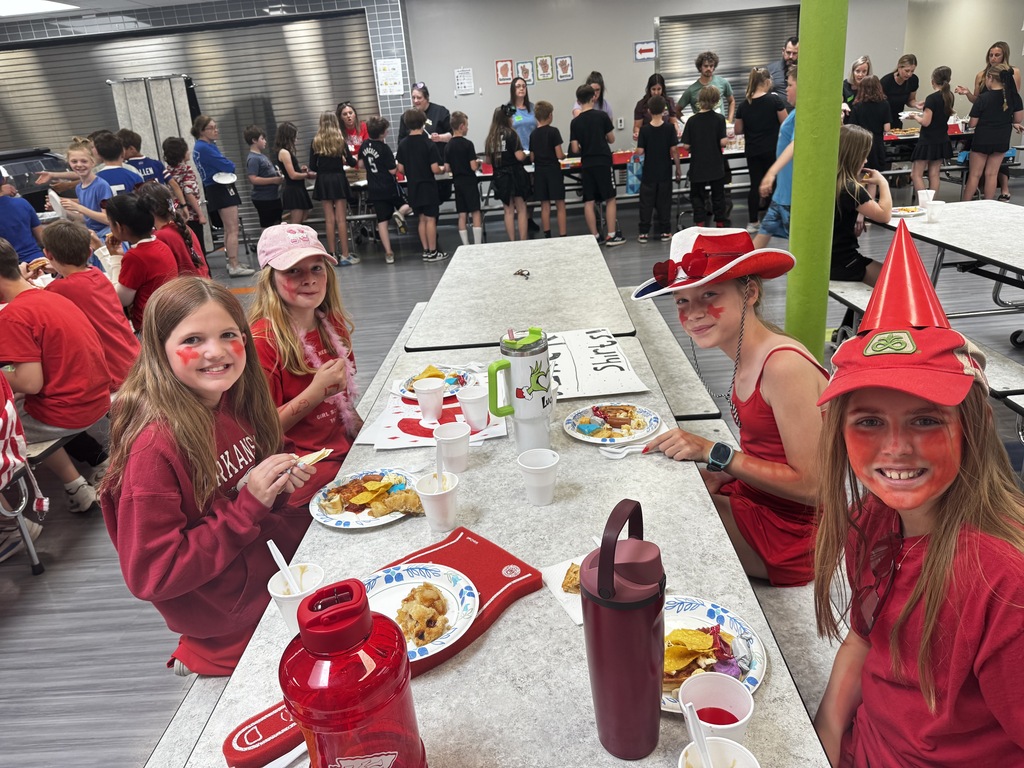 Four girls wearing red shirts, face paint, and spirit hats smile at the camera while sitting at a school cafeteria table during lunch.