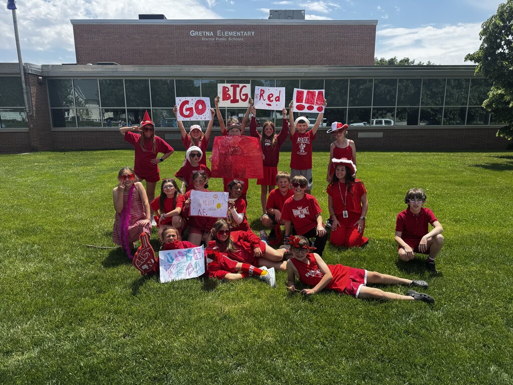 A group of elementary school students dressed in red clothes and face paint pose outside in front of Gretna Elementary School, holding up signs that read "GO BIG RED !!!" and "SUPER RED."