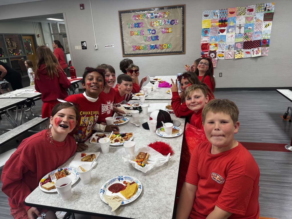 Students with red shirts and red face paint sit along a cafeteria table eating lunch under an indoor bulletin board that reads "Creativity takes courage dare to use your imagination."