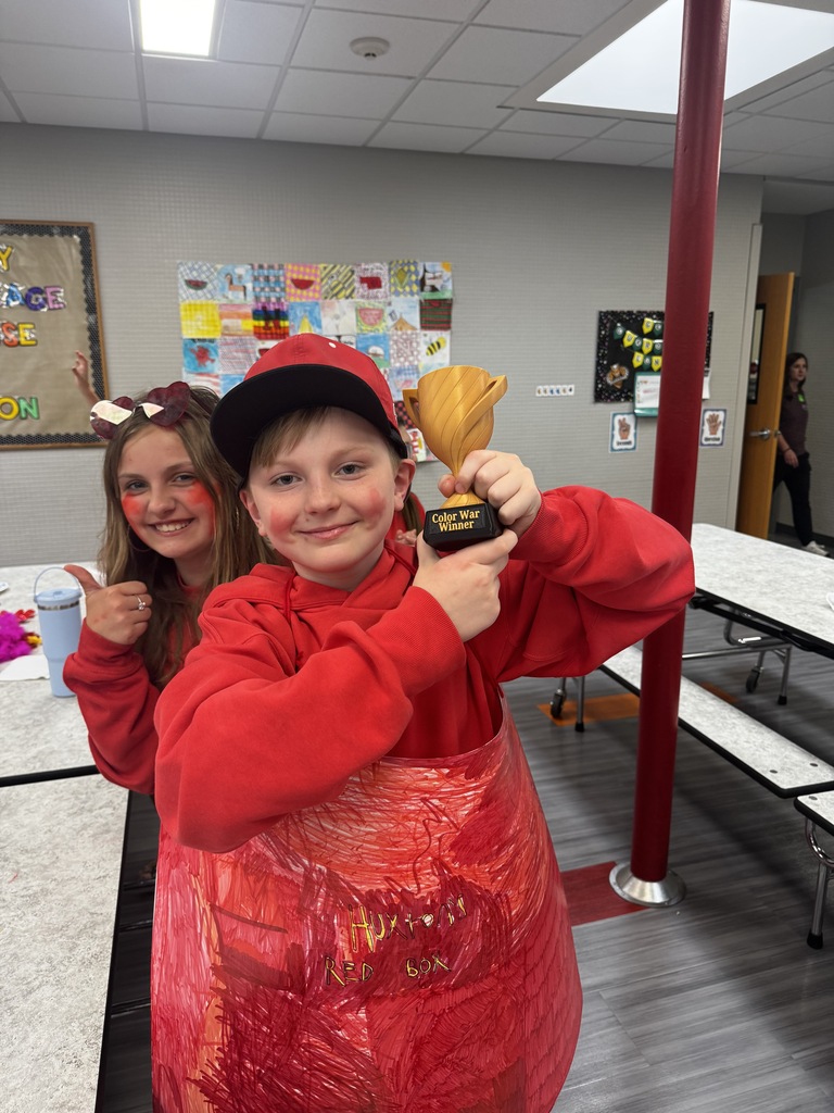 A smiling boy wearing a red sweatshirt and baseball cap proudly holds up a small gold trophy labeled "Color War Winner" next to a girl giving a thumbs-up.