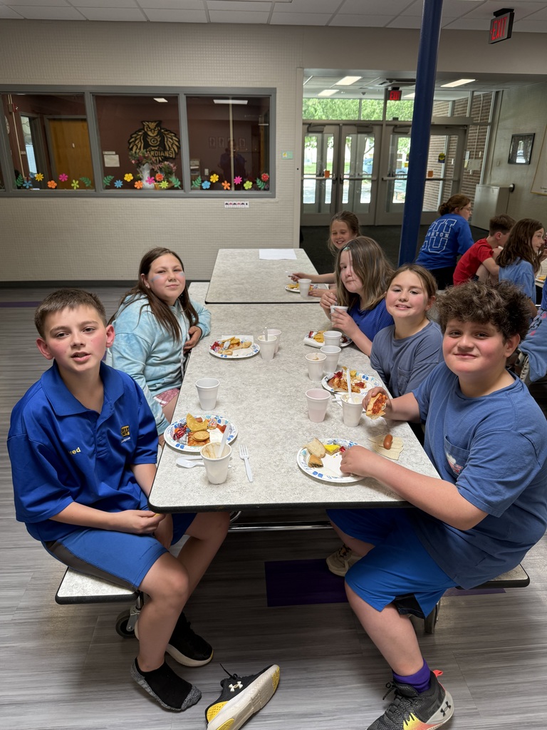 A group of elementary school students wearing blue shirts sit together at a cafeteria table eating pizza, chips, and hot dogs.