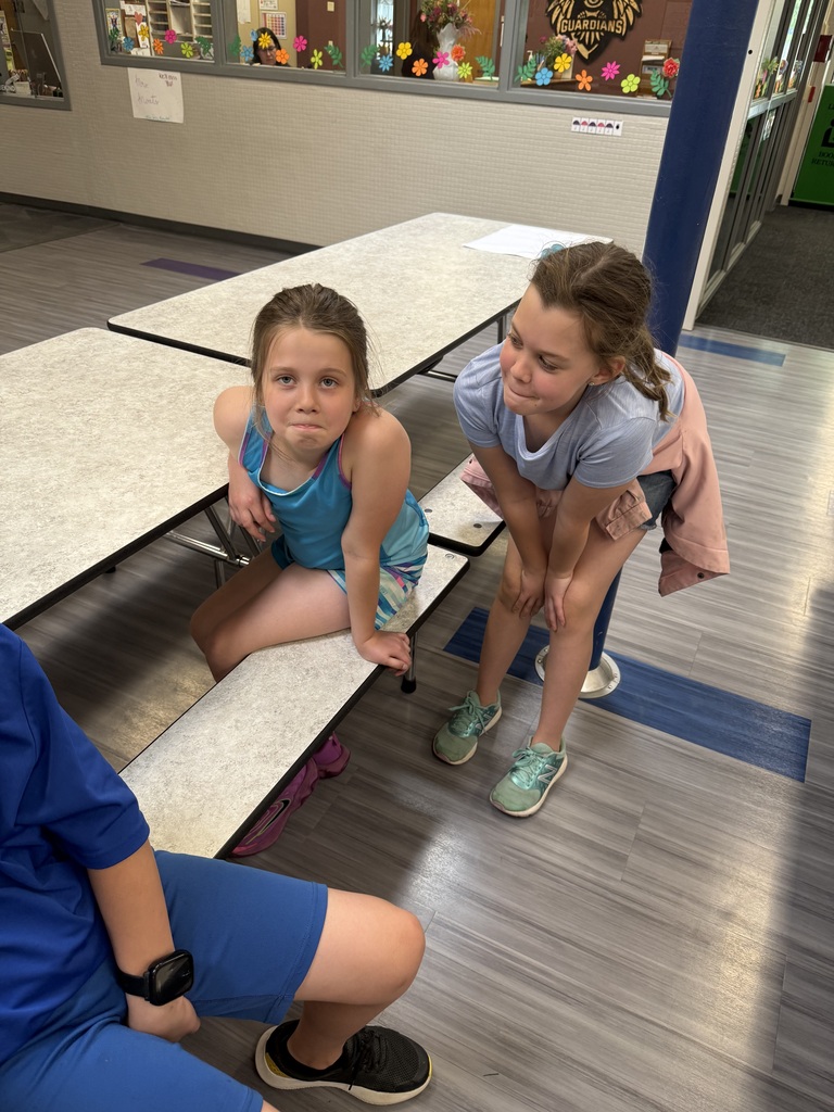 Two young girls, one sitting on a cafeteria bench and one leaning over, look toward the camera with playful, dramatic expressions.