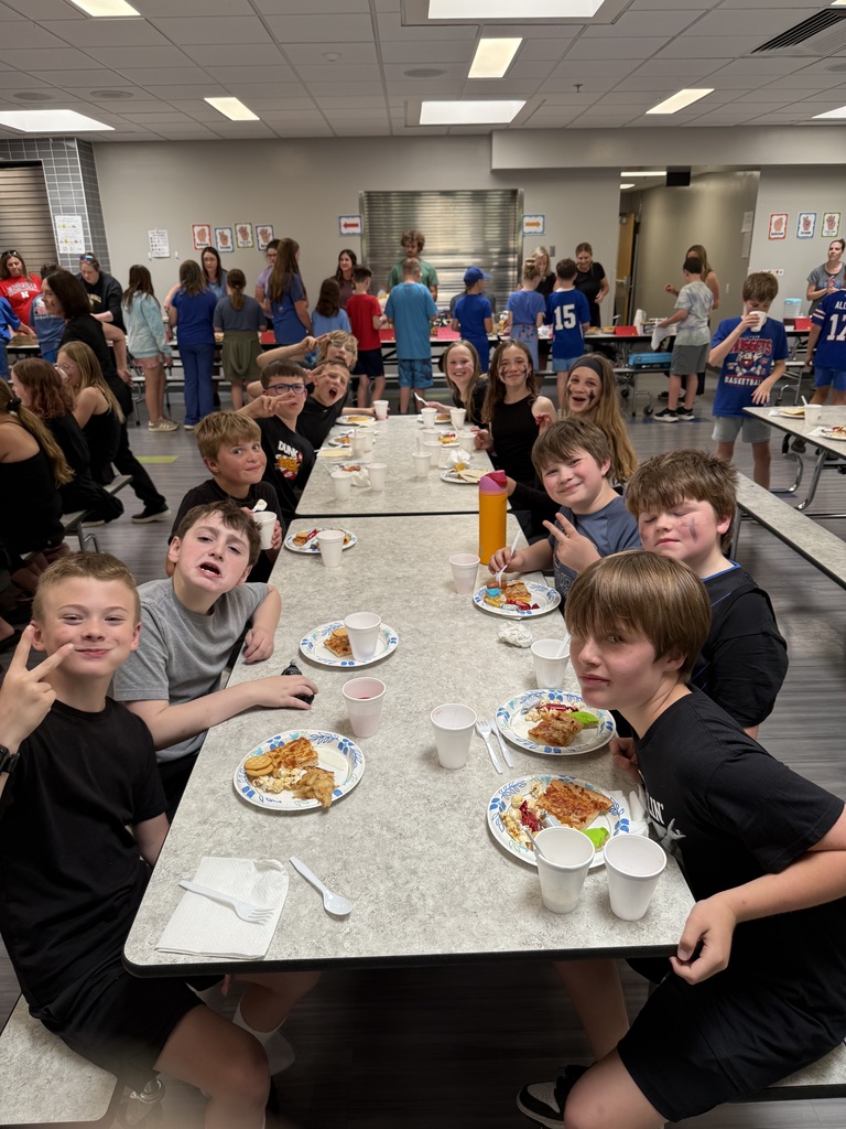 A long cafeteria table filled with boys dressed in black shirts smiling, laughing, and making peace signs at the camera during lunchtime.