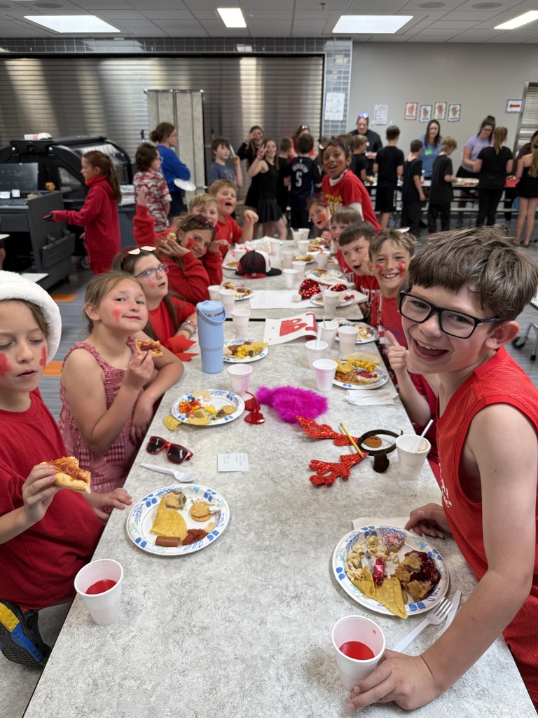 A long table of elementary school students dressed in red and wearing festive holiday accessories smile and make faces while eating lunch in a cafeteria.