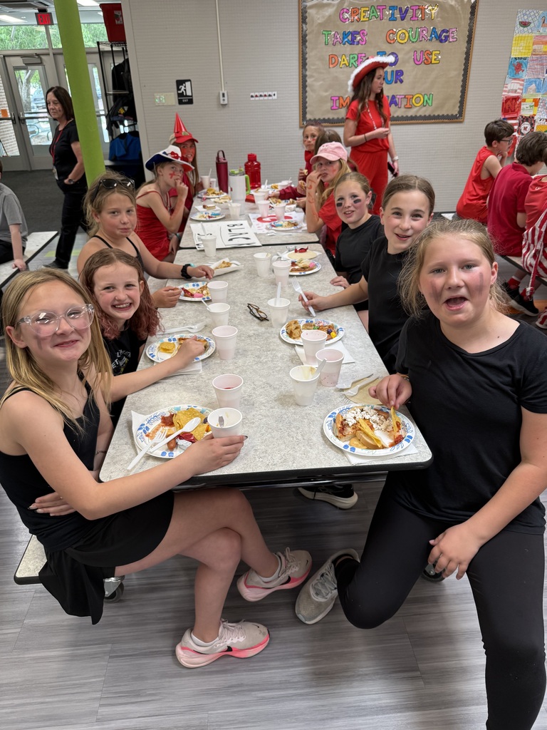 A group of girls wearing matching black tank tops and black face paint sit on one side of a cafeteria table, smiling and eating lunch across from students dressed in red.
