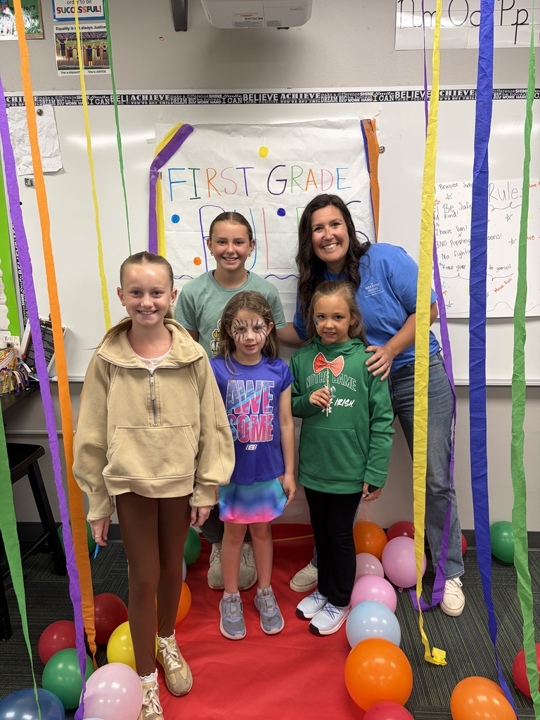 A teacher and four young students smile together inside a classroom on a red runway carpet surrounded by colorful balloons and streamer decorations.