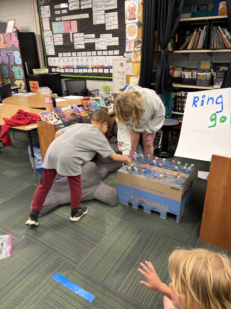 Two children play a homemade "Ring Toss" game constructed out of a large cardboard box topped with colorful lollipops in a classroom setting.