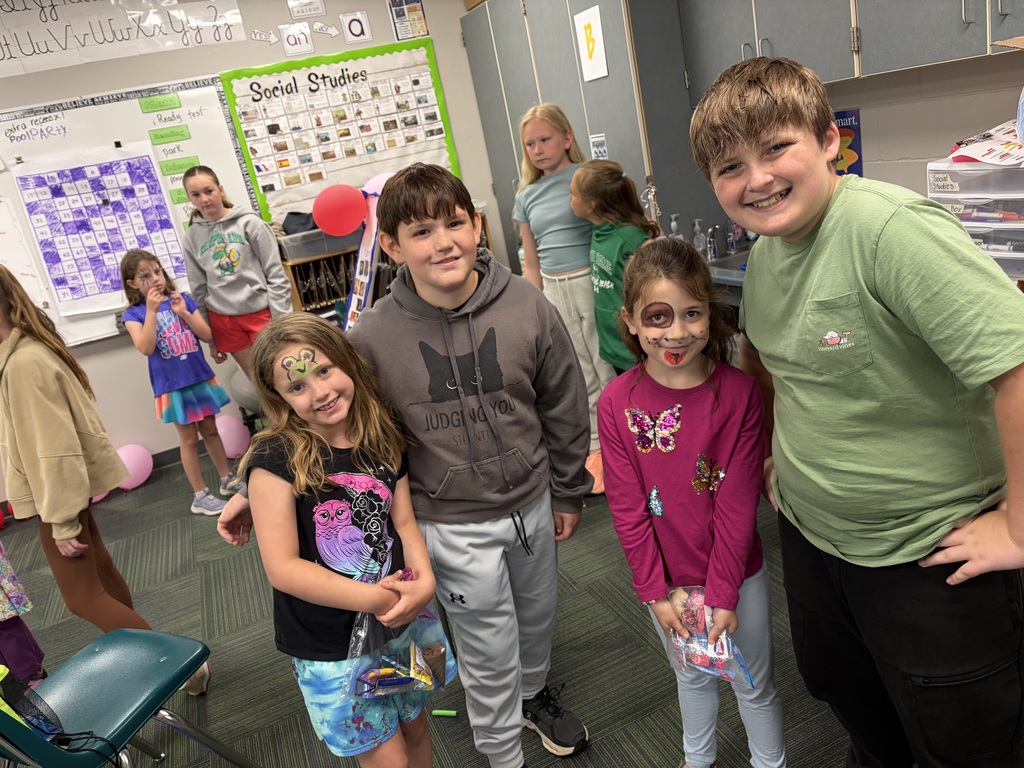 Four students, some with painted faces, smile together in a classroom while holding plastic bags filled with candy prizes.