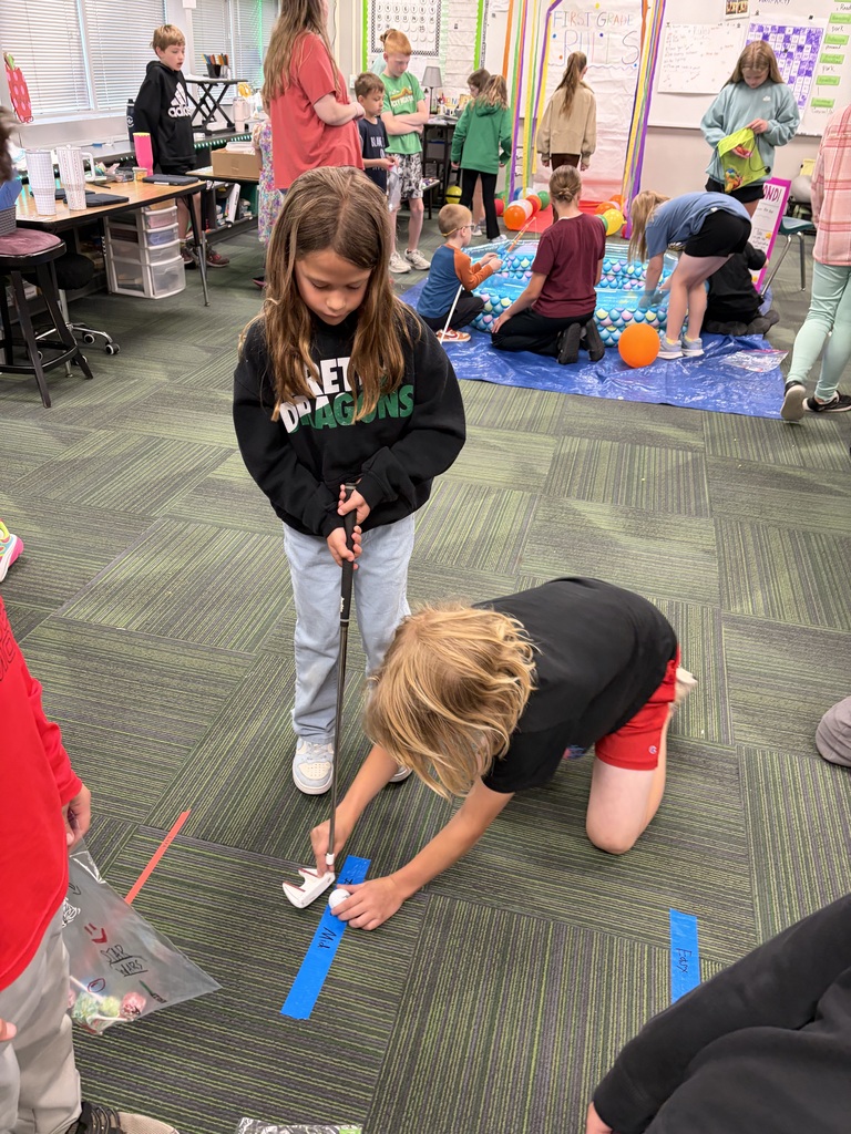 A young girl in a black sweatshirt holds a golf putter while another child on hands and knees helps line up a golf ball on a blue tape line in a busy classroom.