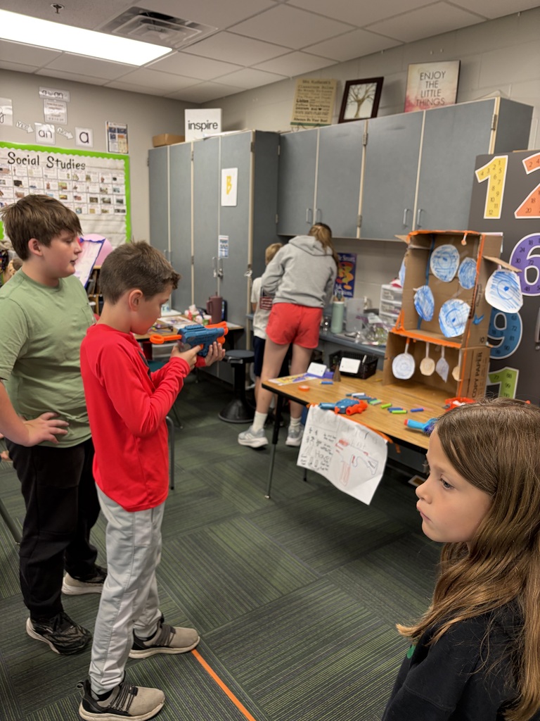 A student in a red long-sleeve shirt aims a blue toy dart blaster at a homemade target game made of paper plates and a cardboard box in a classroom.