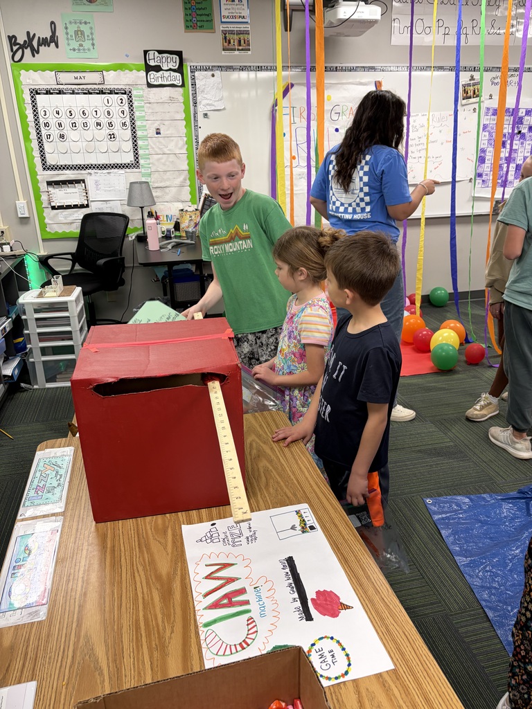 An excited boy in a green shirt demonstrates a large, homemade red cardboard "claw machine" game to two other students standing by a classroom desk.