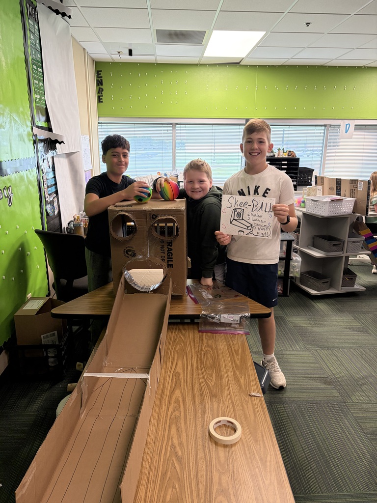 Three smiling boys stand behind a long classroom table featuring a homemade cardboard skee-ball game and holding a hand-drawn instruction sign.