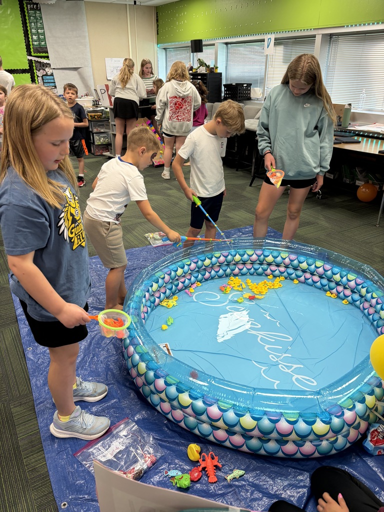 Several elementary students gather around an inflatable, mermaid-patterned pool in a classroom using small nets and plastic fishing poles to catch toy ducks.