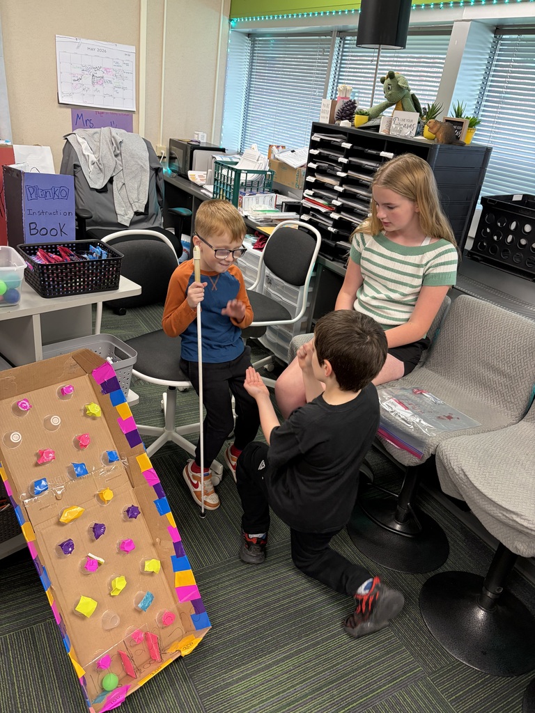 Three students interact next to a colorful, homemade cardboard Plinko game set up on a classroom floor.