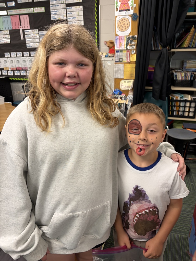 A close-up portrait of a smiling older girl standing next to a younger boy with a puppy dog face paint design in a classroom.