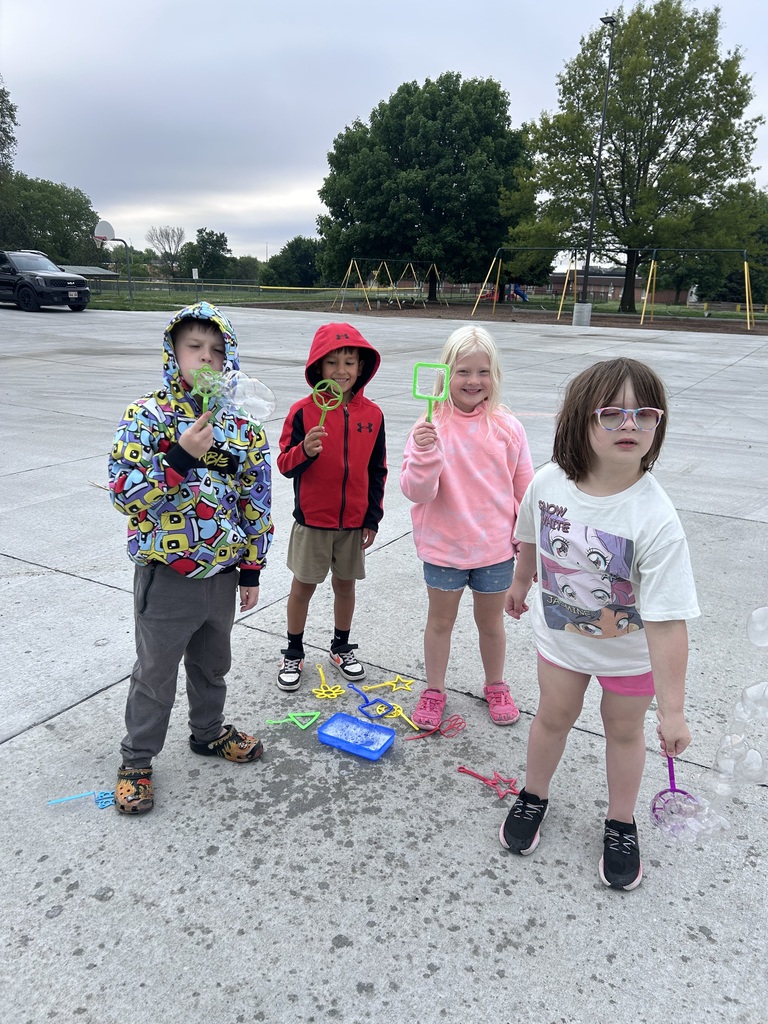 Four young children standing together on a concrete playground holding colorful plastic bubble wands, with playground swings and trees in the background.
