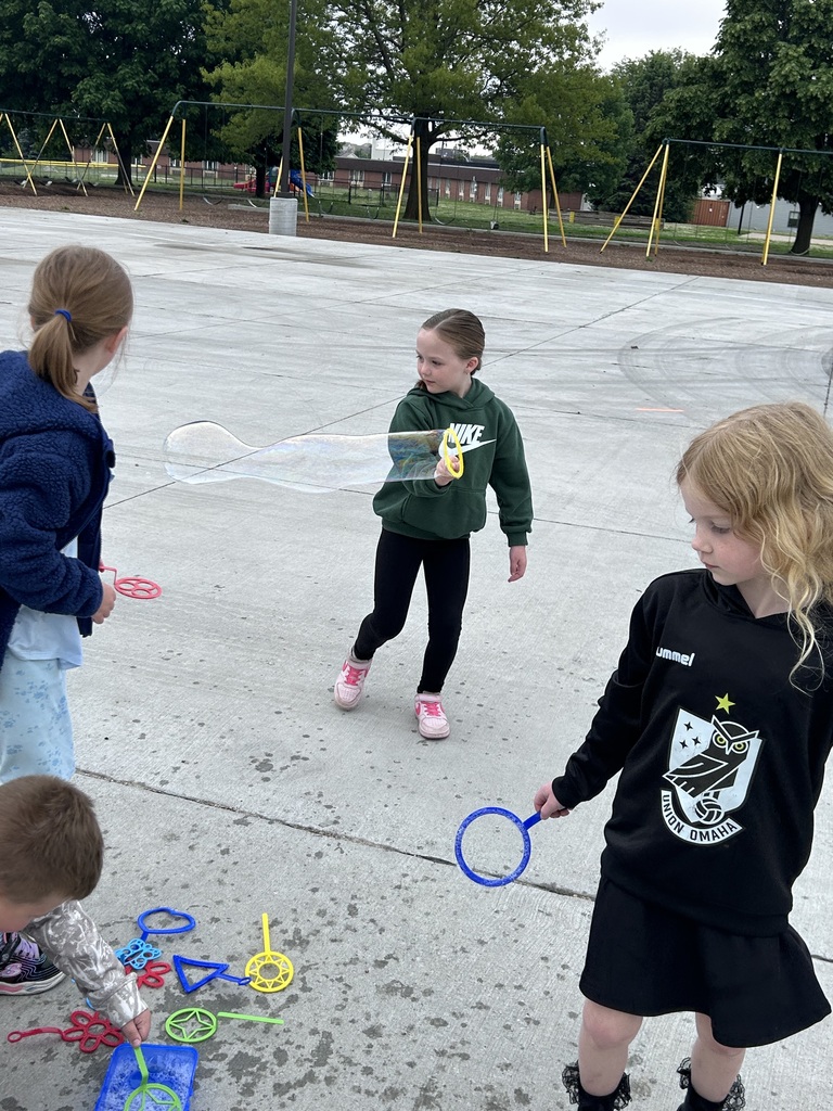 A young girl in a green Nike hoodie creates a long, cylindrical bubble with a yellow wand while other children stand nearby on a concrete playground.