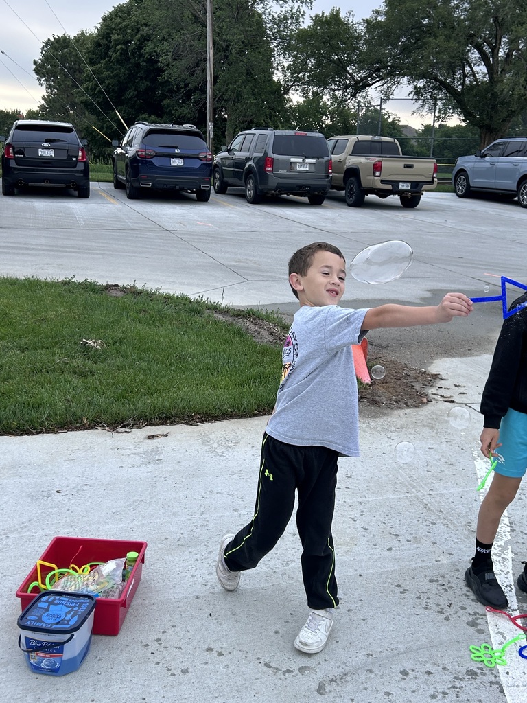 A young boy in a grey shirt and black sweatpants smiles as he waves a blue wand to create a large bubble on an outdoor concrete surface.