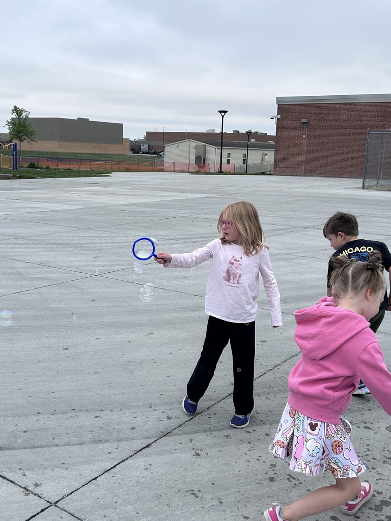 A young girl with pink glasses waves a blue wand to make bubbles on an open concrete playground as other children play around her.