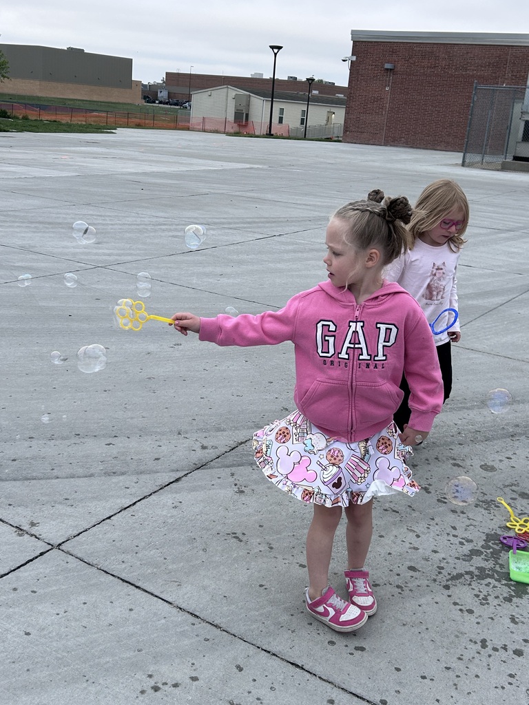 A young girl with her hair in buns, wearing a pink GAP hoodie and a patterned skirt, waves a yellow wand to create a stream of bubbles.