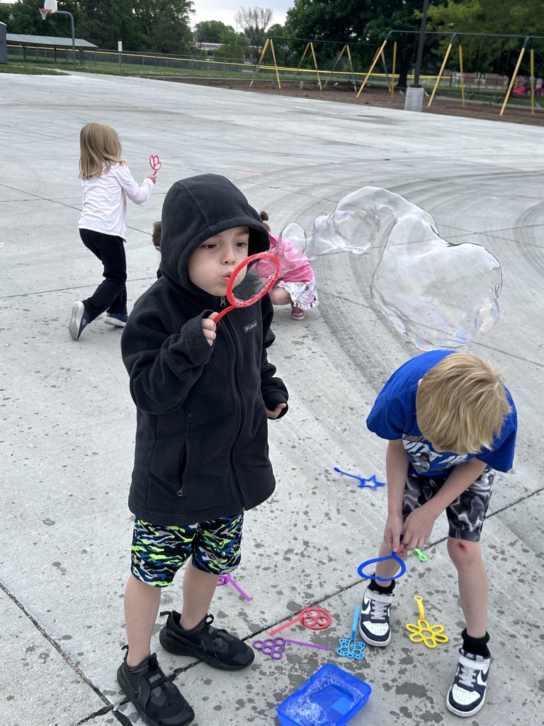 A boy in a black hooded jacket blows a massive, wavy bubble through a red wand while another boy leans over bubble solution nearby.