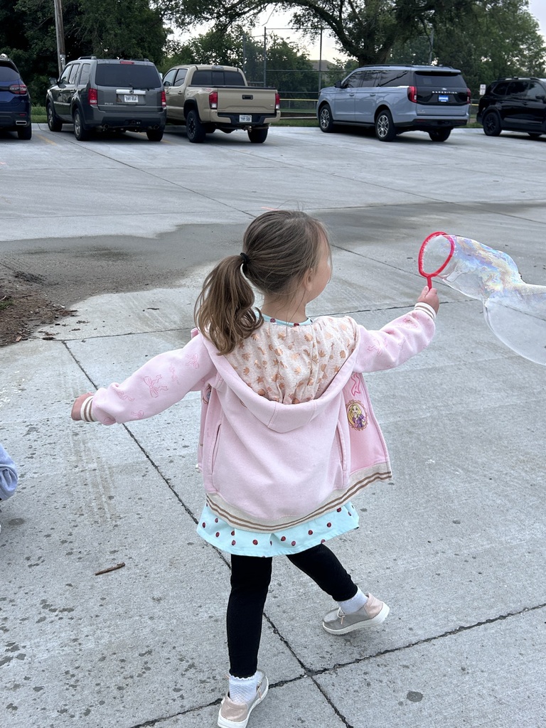 Candid back view of a young girl in a pink jacket and black leggings running with a pink wand, trailing a large bubble behind her on a concrete surface.