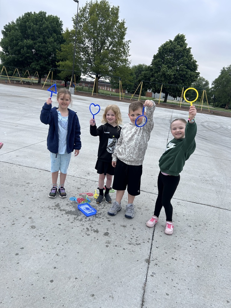 Four smiling children stand in a row on a concrete playground, each holding up a differently shaped colorful bubble wand.