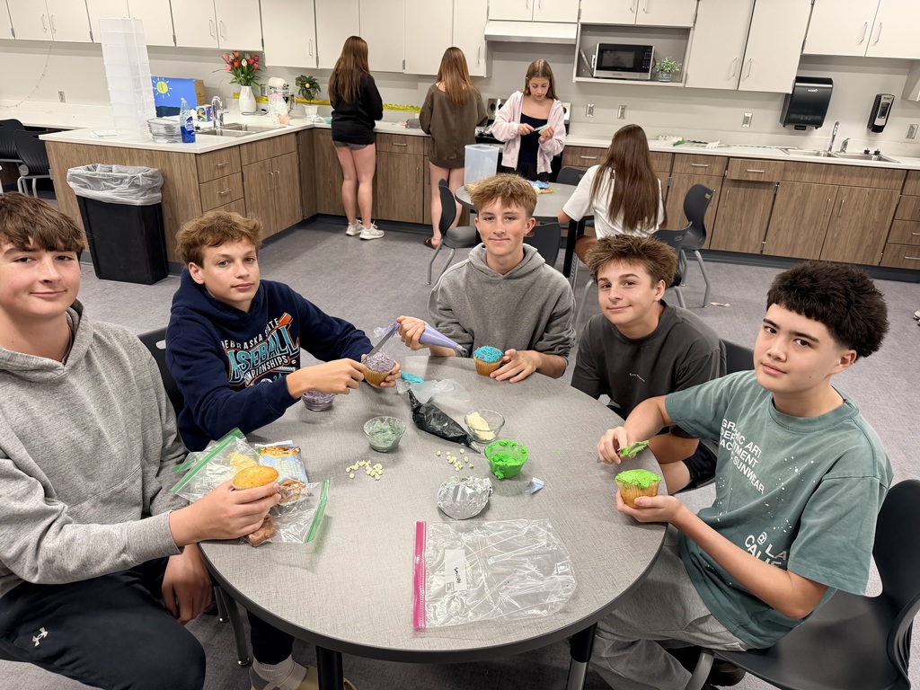 A group of students sit a desk and frost cupcakes. 