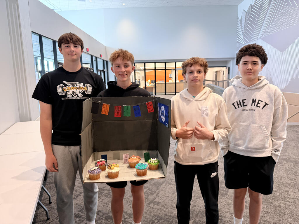 Four boys look at the camera and smile. One is holding a trifold box cutout with cupcakes on the bottom. 