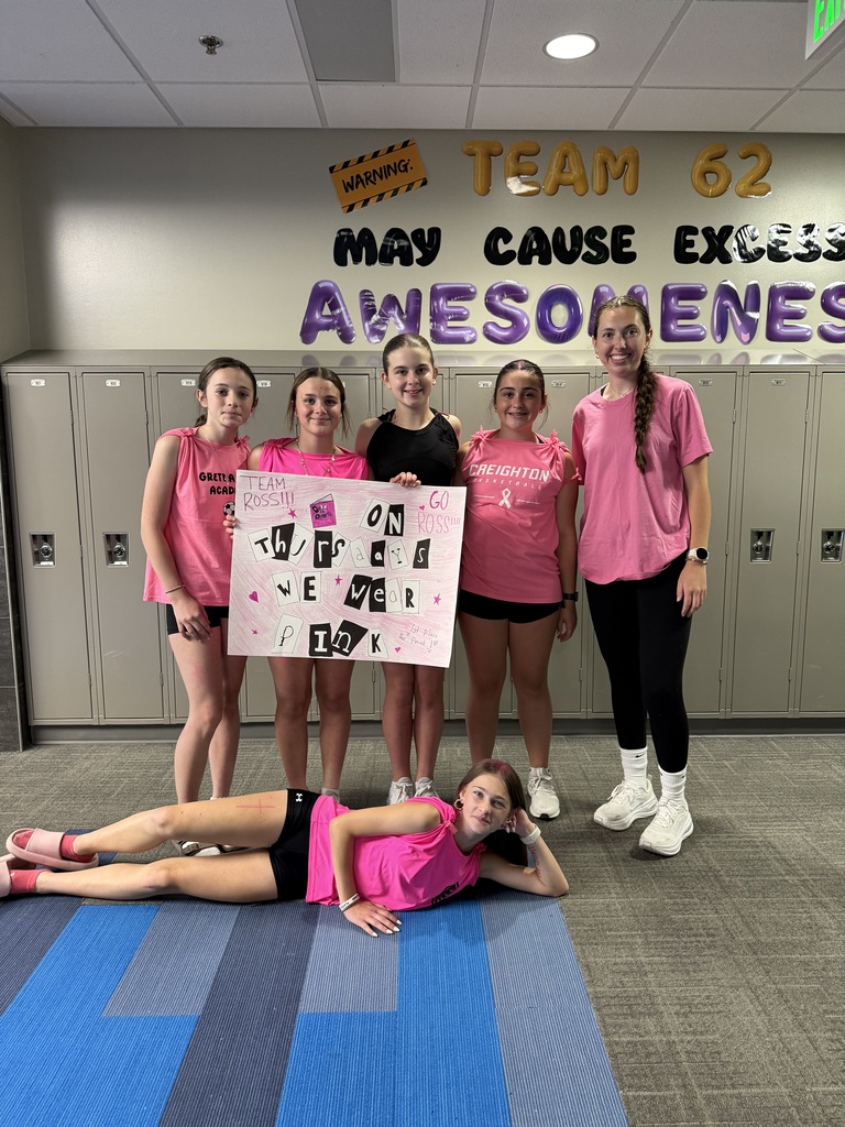 Photo with teacher and four students standing wearing their pink outfits for kickball. They are holding a sign that says, "On Tuesdays, we wear pink," and there is one more student laying on the ground in front of them.