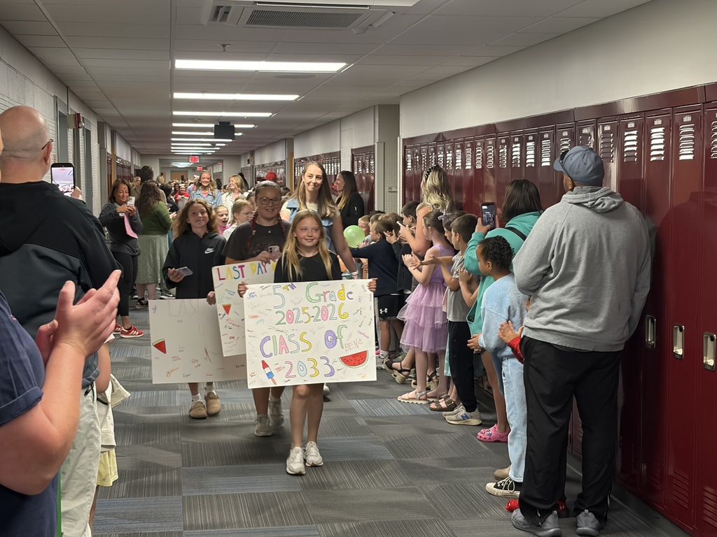 A school hallway filled with people celebrating the last day of school. A young girl in the foreground holds a large white sign that reads "5th Grade 2025-2026, Class of 2033" decorated with drawings of a watermelon slice and a popsicle.