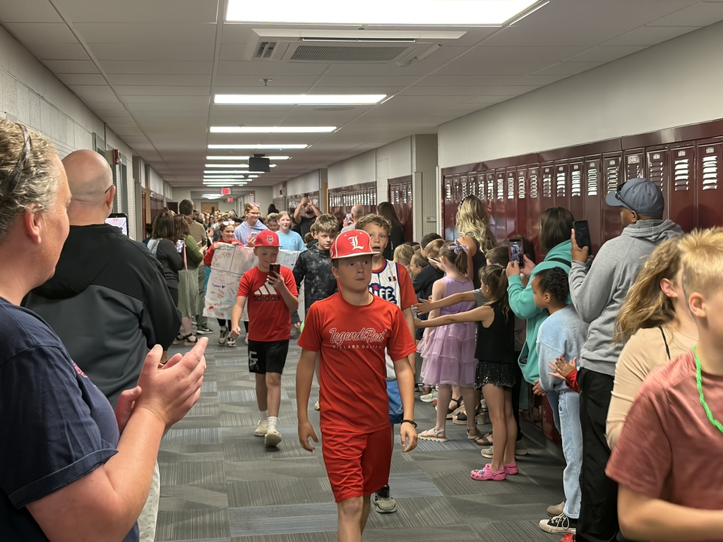 Students walking down a school hallway during a last-day-of-school celebration. Two boys in matching bright red t-shirts and baseball caps walk in the center, while parents and staff line the sides to watch and applaud.