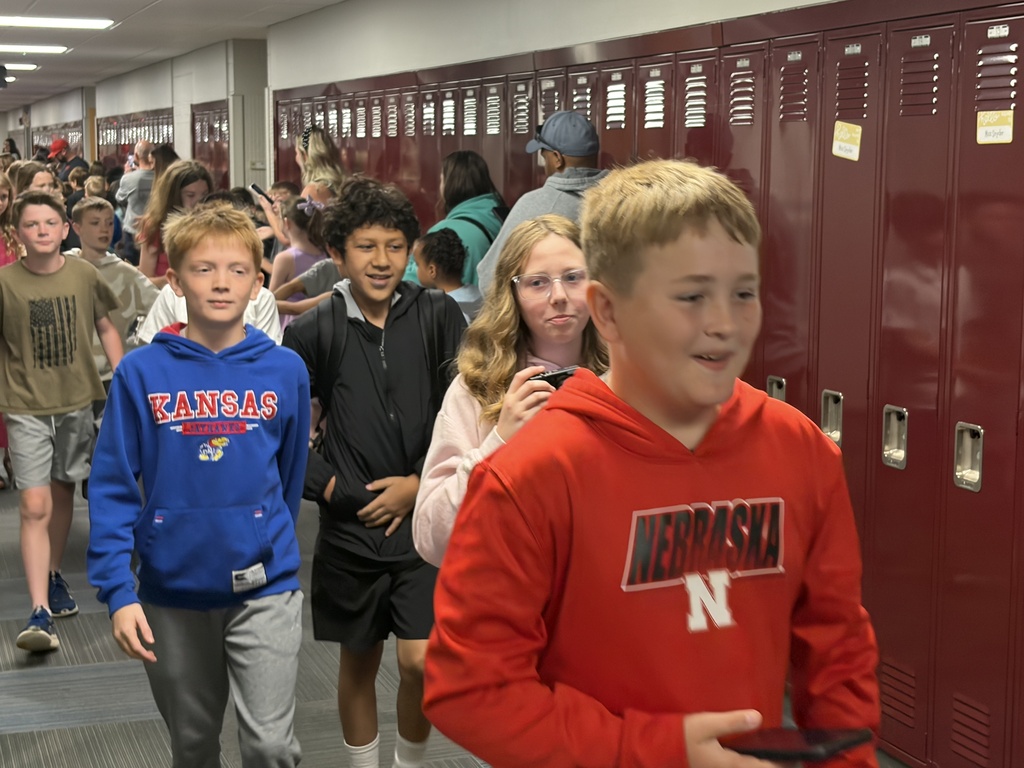 A group of students walking down a school hallway lined with maroon lockers. A boy in the foreground wears a red Nebraska hoodie, while another boy close behind wears a blue Kansas hoodie.