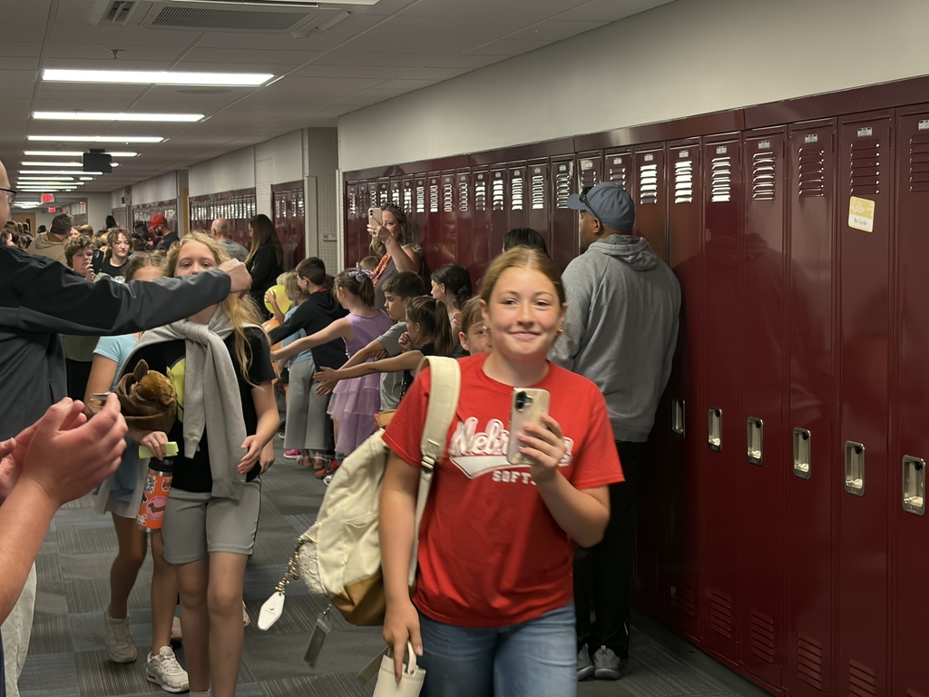 A smiling girl in a red Nebraska t-shirt and backpack walks toward the camera in a crowded school hallway. Behind her, younger students line up against the lockers to high-five the passing older students.