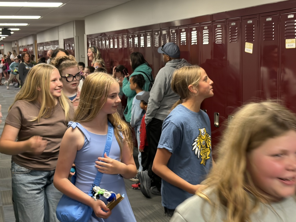 A group of girls smiling and walking down a school hallway past a line of maroon lockers. One girl wears a light blue dress with a matching crossbody bag and holds a small trophy.
