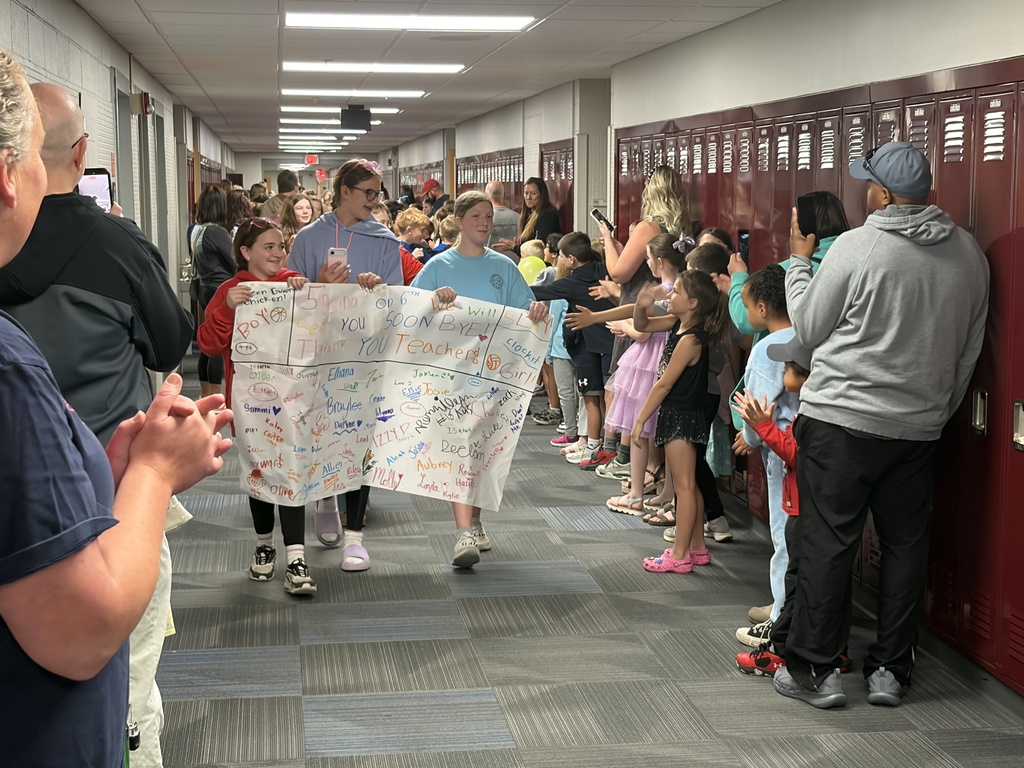 Three young girls walking down a school hallway holding up a large white banner covered in signatures and hand-drawn doodles. The banner reads "5th Grade on 6th, We will miss you, SOON BYE!, Thank You Teachers!"