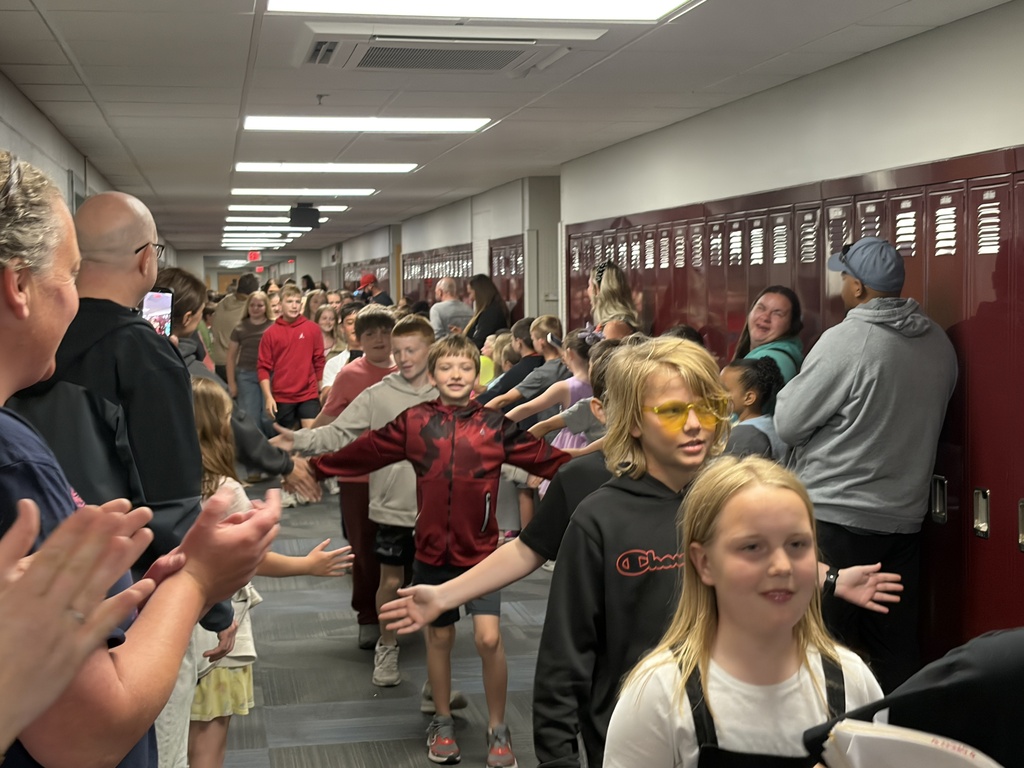 Students walking down a school hallway with their arms outstretched to high-five onlookers. A boy in the foreground wears a black hoodie and bright yellow-tinted sunglasses.