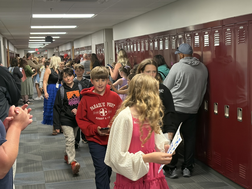 Students walking in a line down a school hallway past onlookers and maroon lockers. A girl with long, wavy blonde hair in a pink dress and white cardigan stands in the foreground looking down at a flyer.
