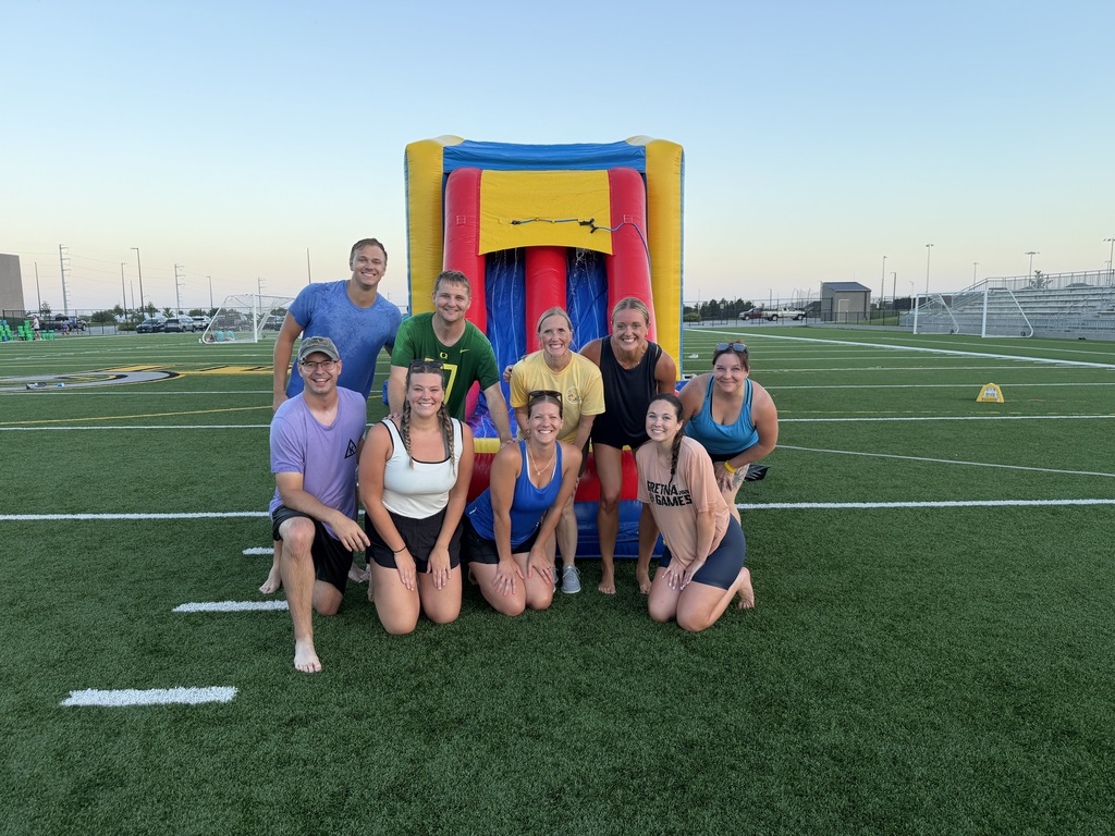 A group of nine smiling adults in casual athletic wear posing together on a green turf field in front of a colorful, wet inflatable water slide.
