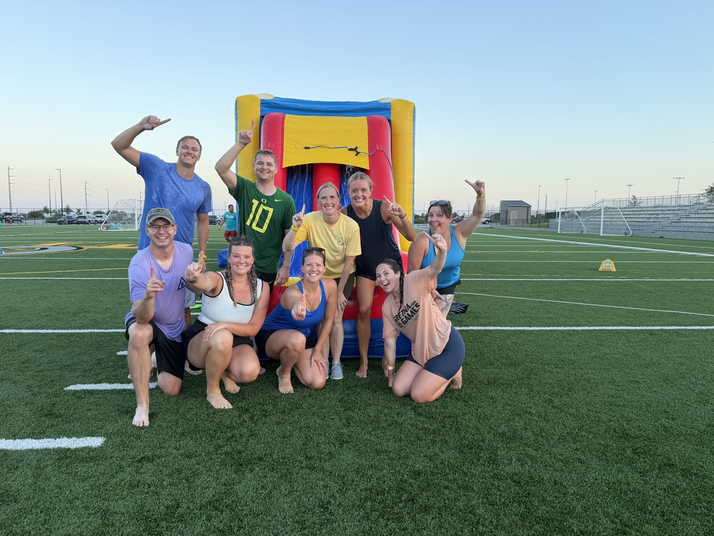 A group of nine adults posing cheerfully on a sports turf field in front of an inflatable water slide, with several individuals holding up their index fingers to signal "number one."