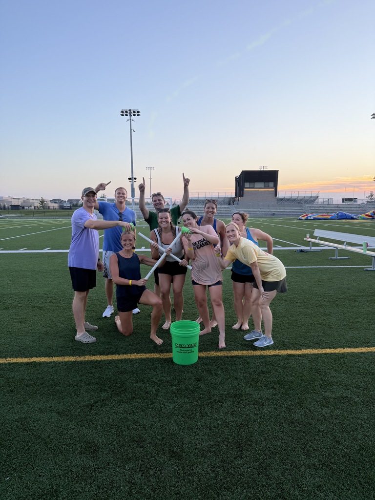 A group of nine people posing playfully on a turf field around a bright green Menards bucket, holding white PVC pipes and plastic cups during an outdoor game at dusk.
