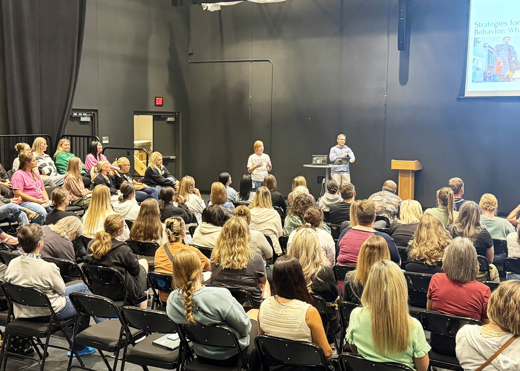 a large group of staff members sitting in the black box theater room listening to a presenter at the front of the room
