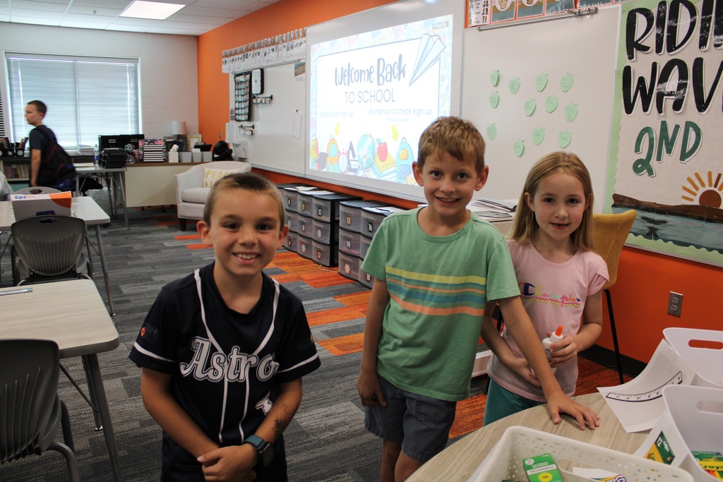 Two boys and a girl stand behind a table smiling at back to school night.
