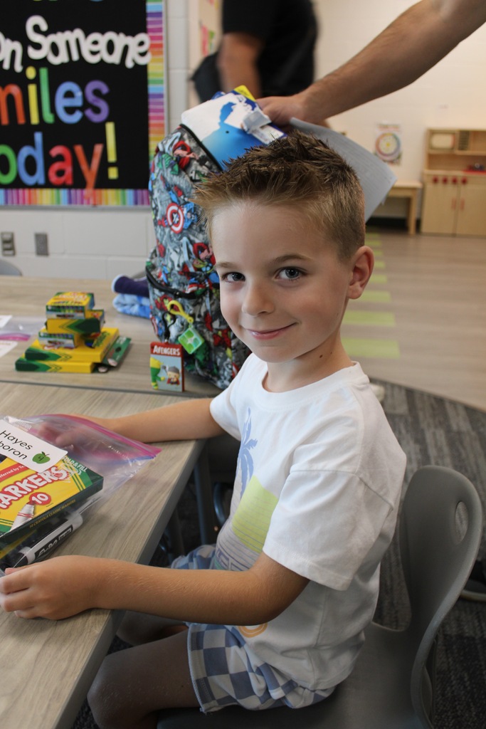 A new kindergarten boy sits at a table with a bag of school supplies.