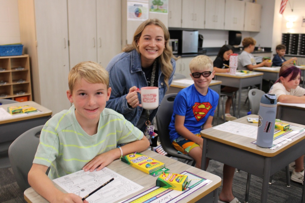 Two fourth grade boys sit at desks with Miss Clark smiling behind them.