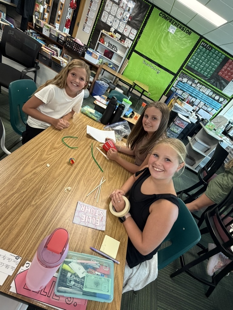 Three girls smiling at a desk with school supplies, tape, and project materials.