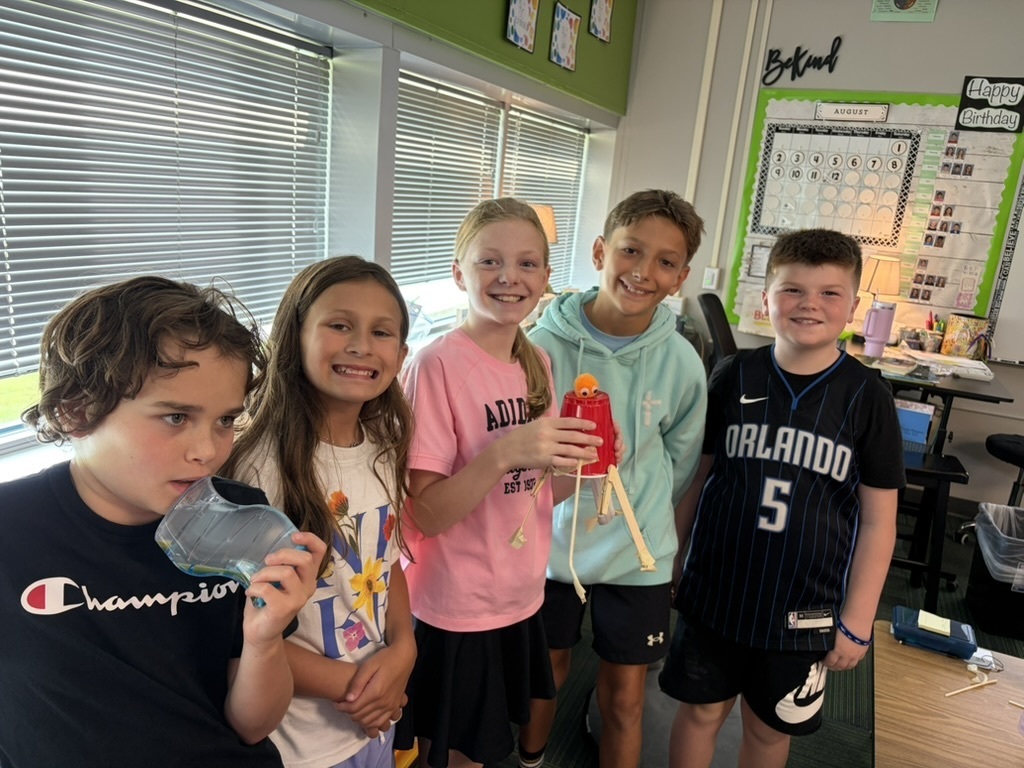 Five students standing in a classroom smiling as one girl holds up a red plastic cup tower project.